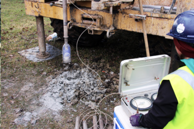 Image of worker in safety gear and equipment, drilling for samples as part of a field investigation.