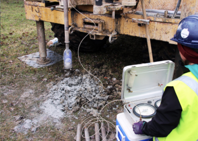 Image of worker in safety gear and equipment, drilling for samples as part of a field investigation.