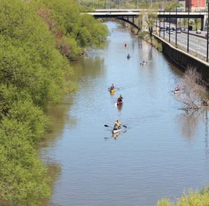 Image of several groups of people canoeing on a river with greenery on the left, and a highway on the right.