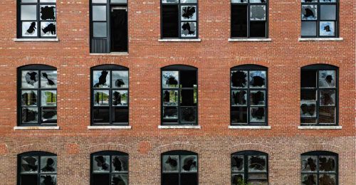 Face of brick abandoned factory building with broken windows and overgrowth.