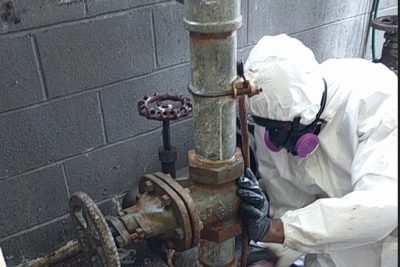 Image of a worker in hazmat gear, taking a close look at a building pipe.