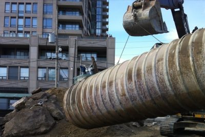 Urban decommissioning site with large equipment moving a pipe and an apartment building in the background.