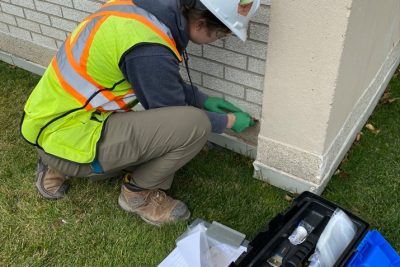 Image of worker in safety gear taking samples from a brick building, with a tool box and clipboard beside them.